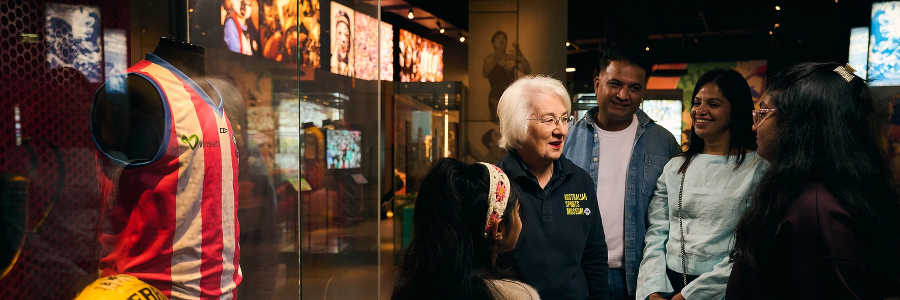 An Australian Sports Museum staff member leading a group of visitors past a football display