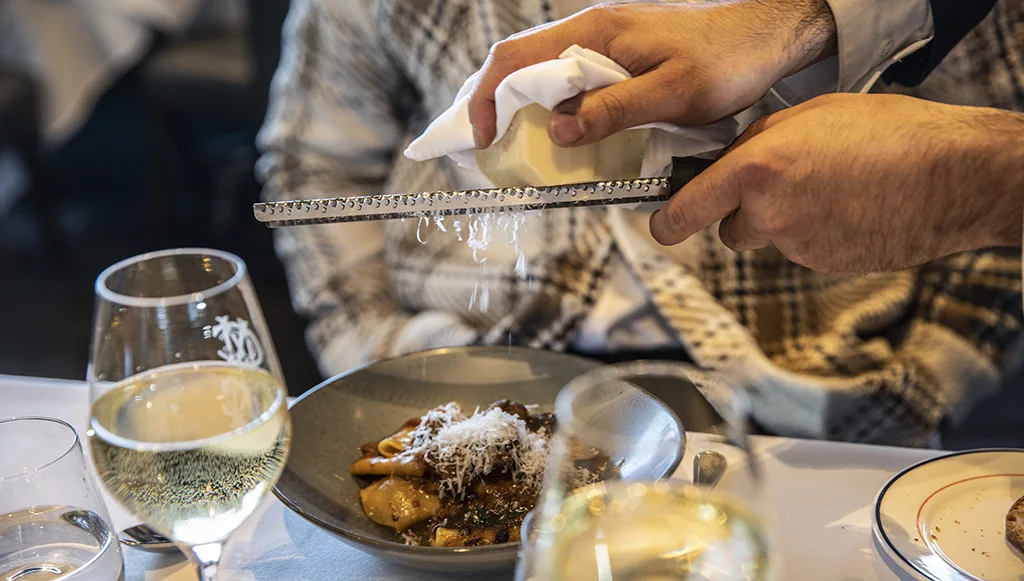 A waiter serves cheese with a pasta dish. Glasses of white wine sit on the white tablecloth.