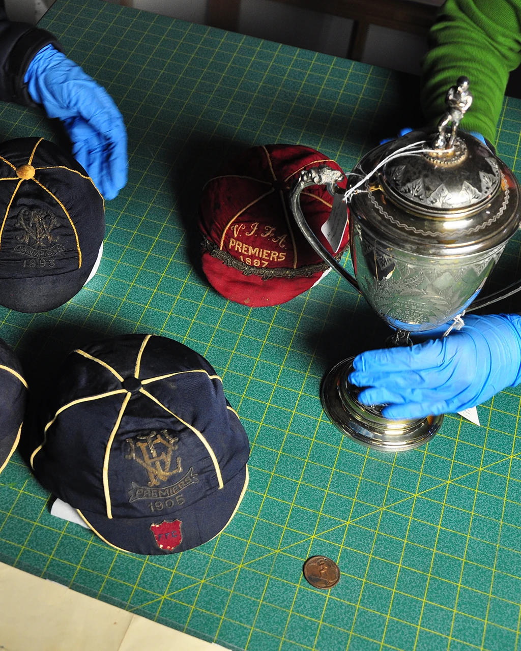 Gloved hands handling historic football caps, including a VFA Premiers 1897 cap, alongside a silver trophy