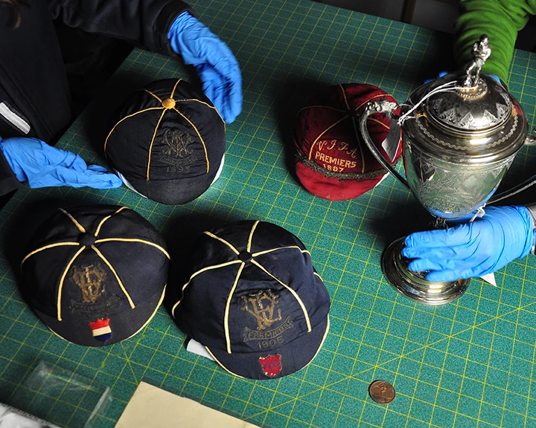 Gloved hands handling historic football caps, including a VFA Premiers 1897 cap, alongside a silver trophy