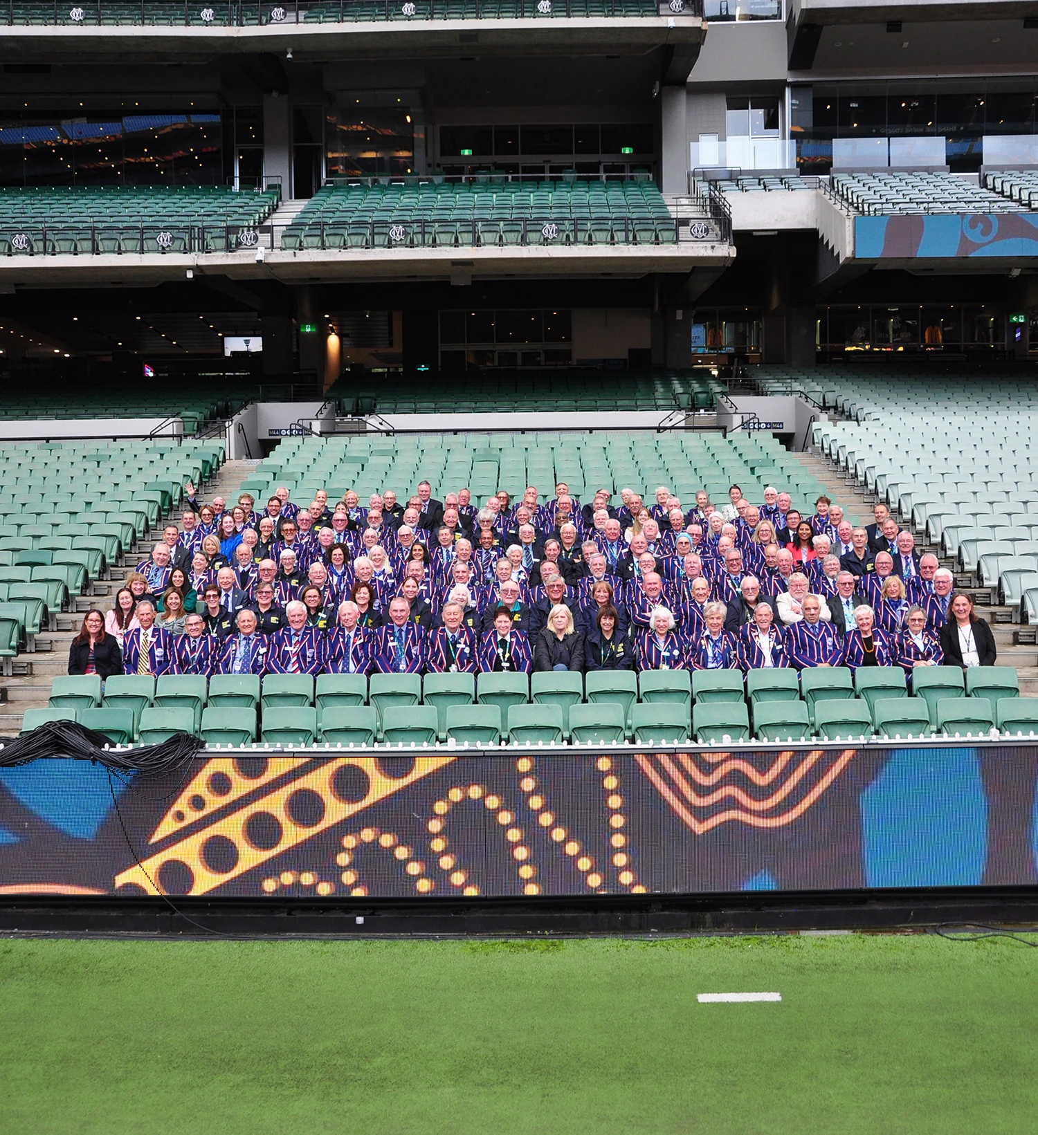 Approx 100 people in MCC volunteer blazers sit in MCG stadium seats together