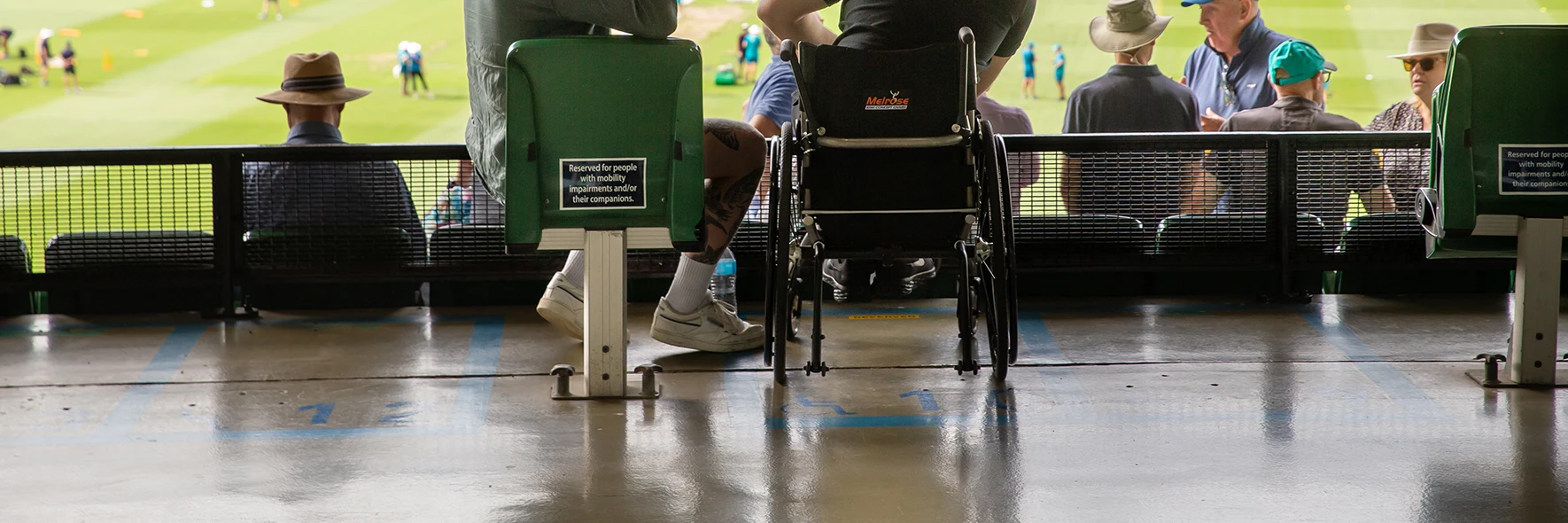 Accessible seating at the back of a stadium section. A person using a wheelchair is next to a companion in a reserved seat.
