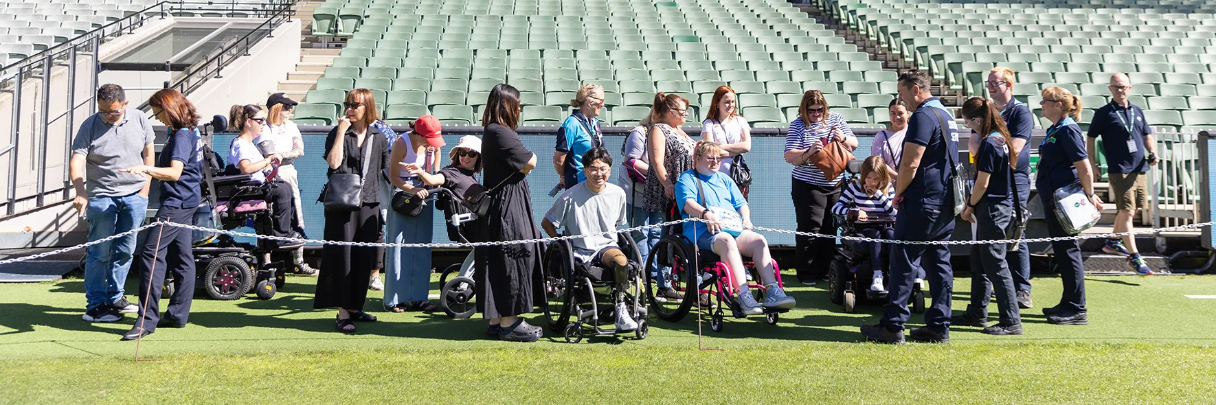 A group tours an empty stadium along the boundary line with some group members using wheelchairs and mobility aides.
