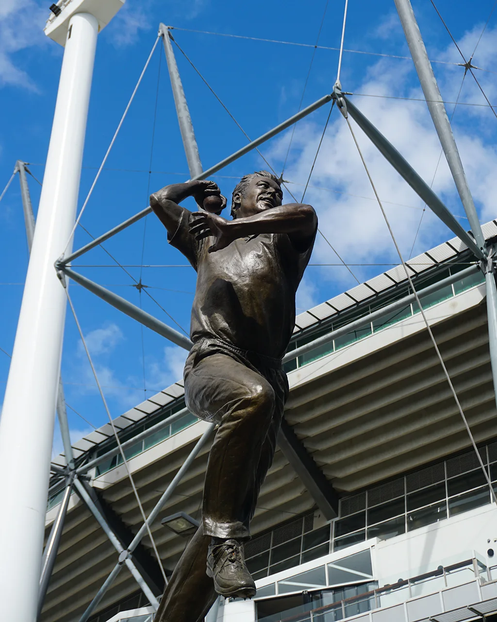 Bronze statue of Shane Warne bowling, on a backdrop of the MCG exterior.