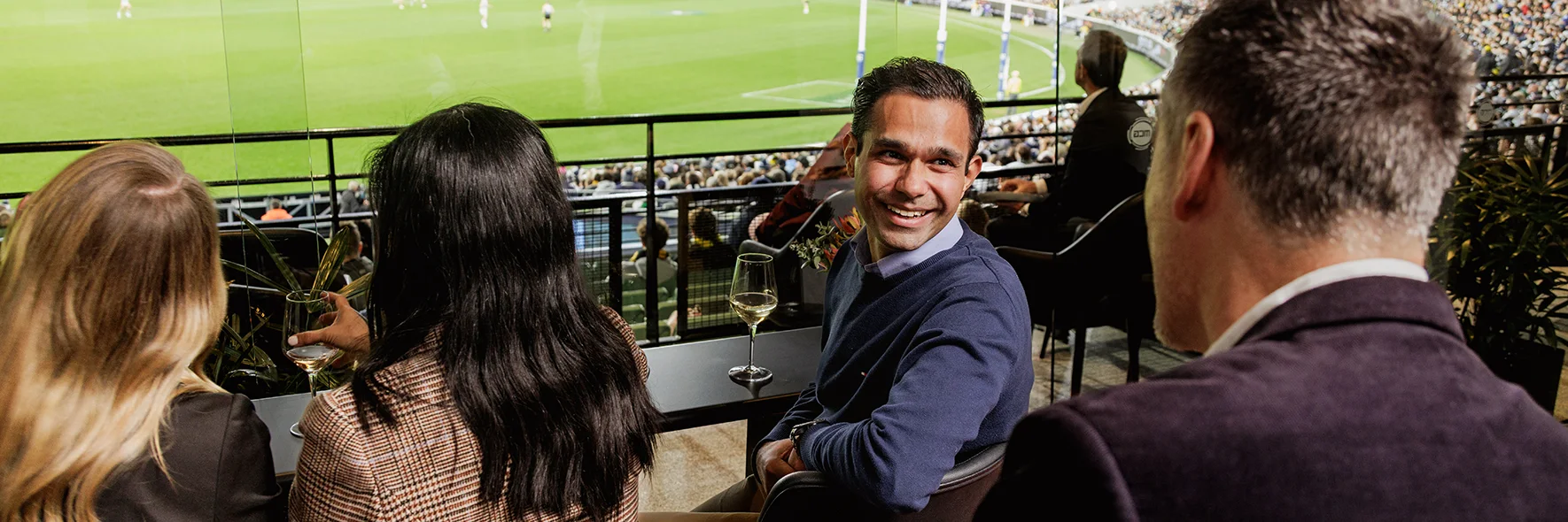 Happy guests enjoying MCG AFL game in private lounge area with wine and goal view.