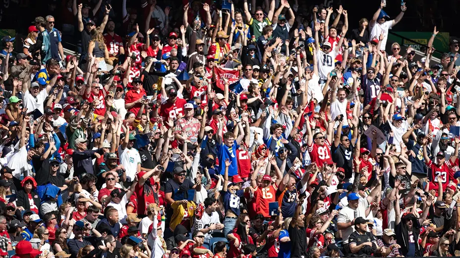 Fans in NFL team jerseys stand with arms raised, celebrating in the MCG stands